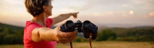 istockphoto-1452691834-612×612 Copy space shot of mature woman giving you binoculars and pointing in the distance, to where to look at while standing on the viewpoint.